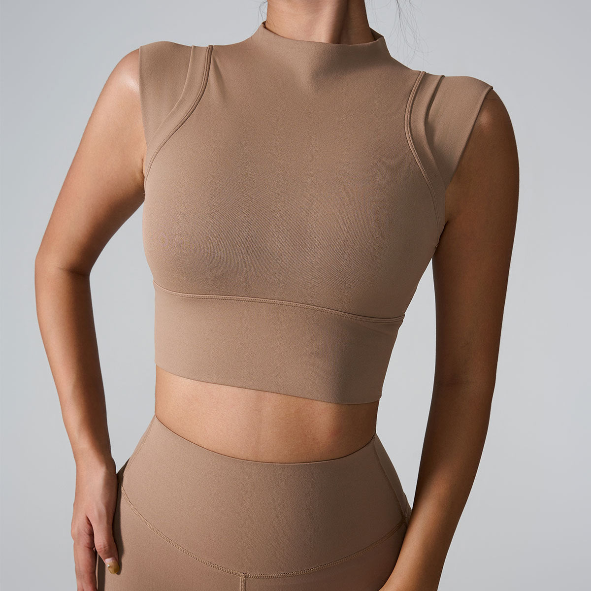 A woman is wearing a taupe seamless mock neck crop top with contoured seam details, paired with matching leggings against gray background