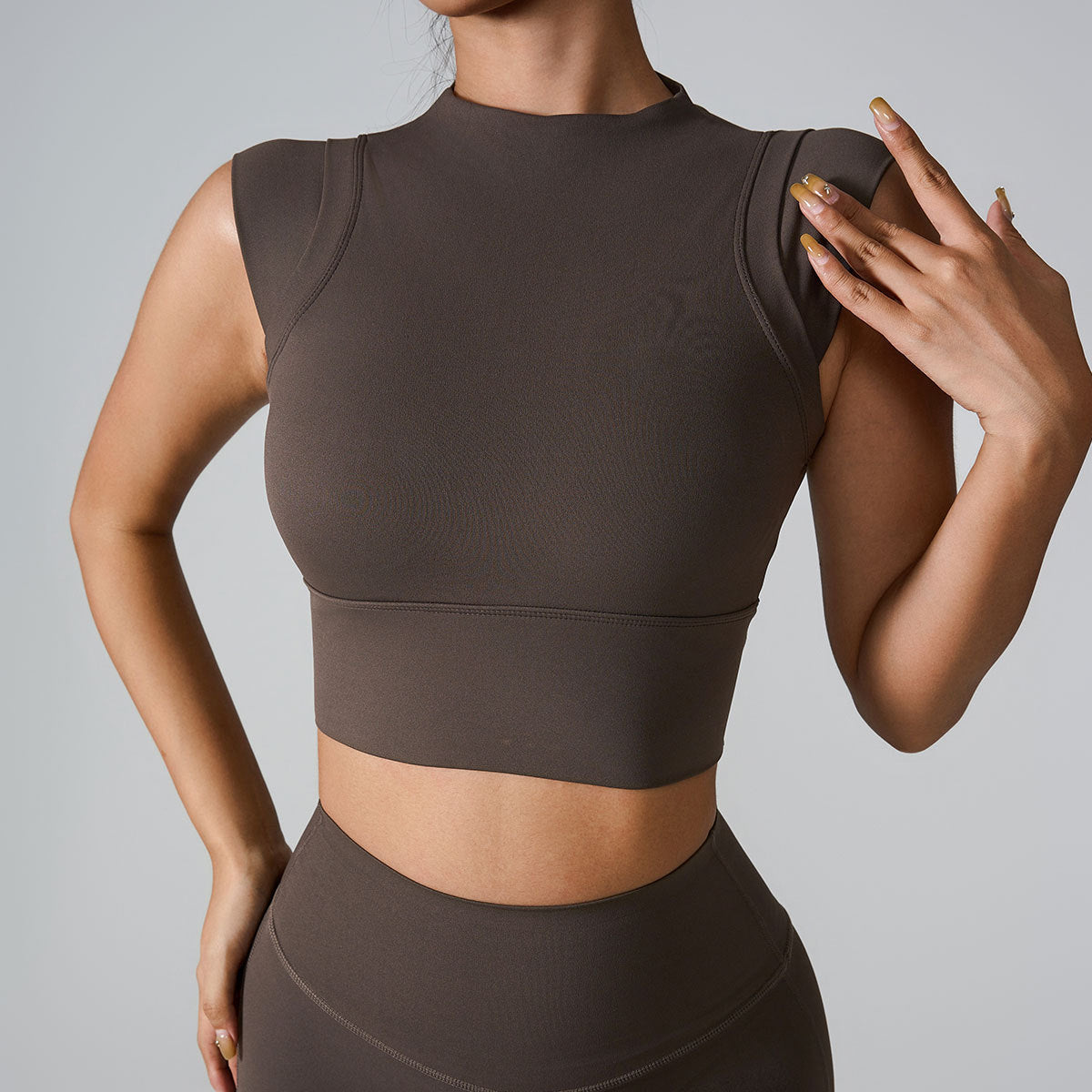 A woman is wearing a chocolate brown seamless athletic crop top with mock neck, touching her shoulder against a light gray backdrop