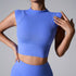 A woman is wearing a periwinkle blue seamless athletic crop top with mock neck, touching the shoulder seam detail against gray backdrop