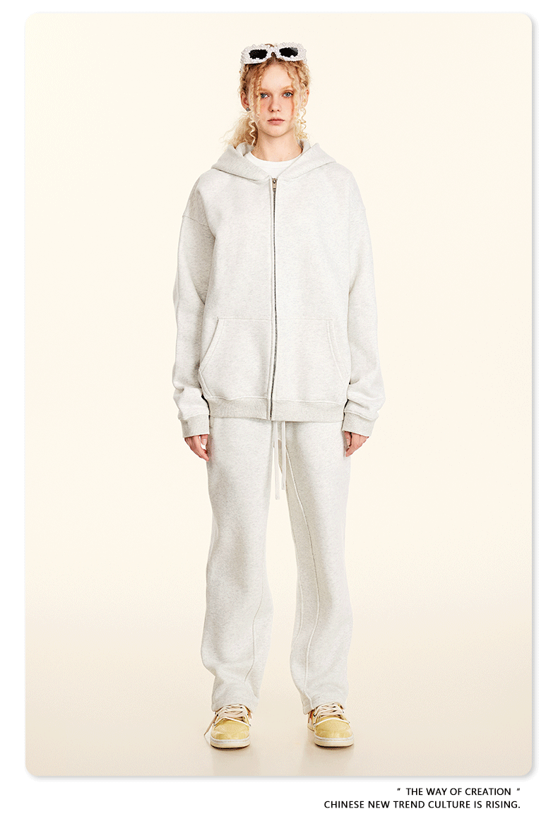Blonde model wearing white oversized zip-up hoodie and matching sweatpants with white sunglasses on head and yellow shoes against cream background