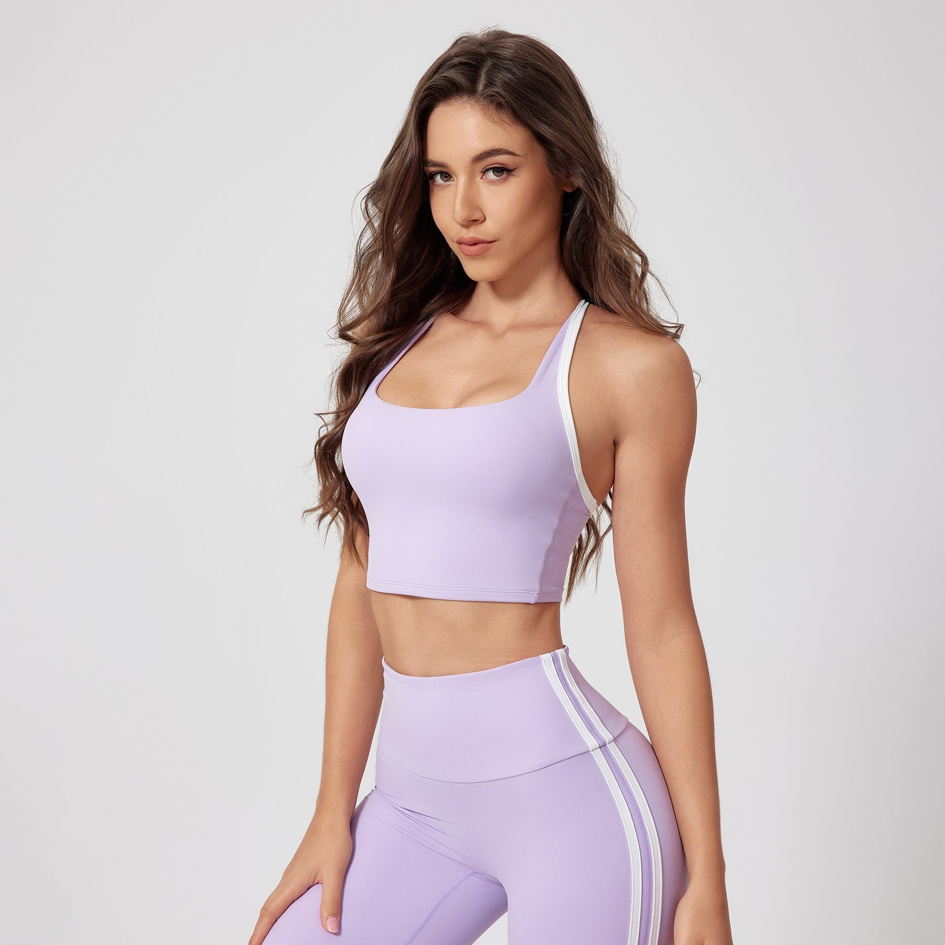 A woman is posing in a lavender sports bra with white straps and matching high-waisted leggings with side stripes against a white backdrop