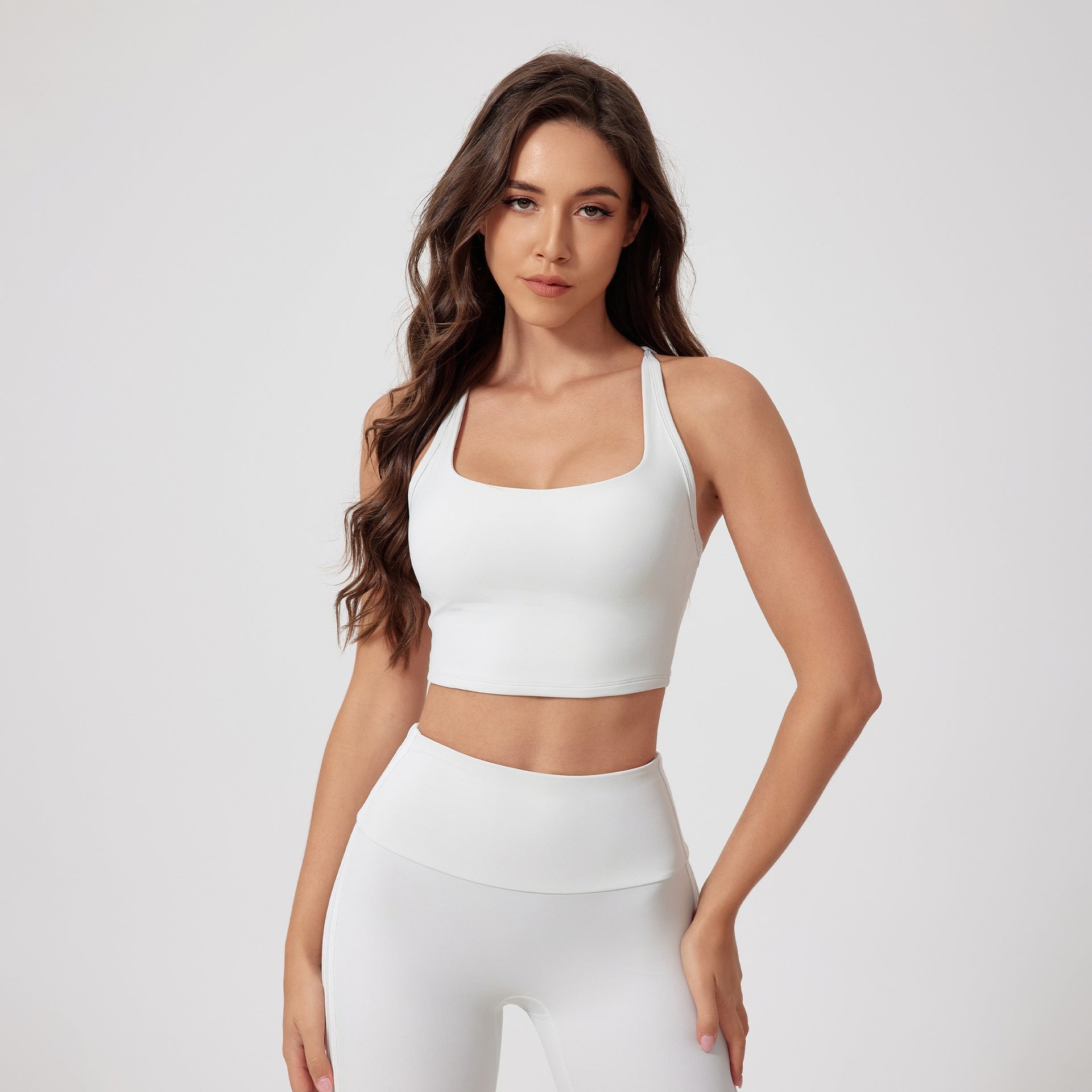 A woman is standing in a white racerback sports bra and matching high-waisted compression leggings against a neutral background