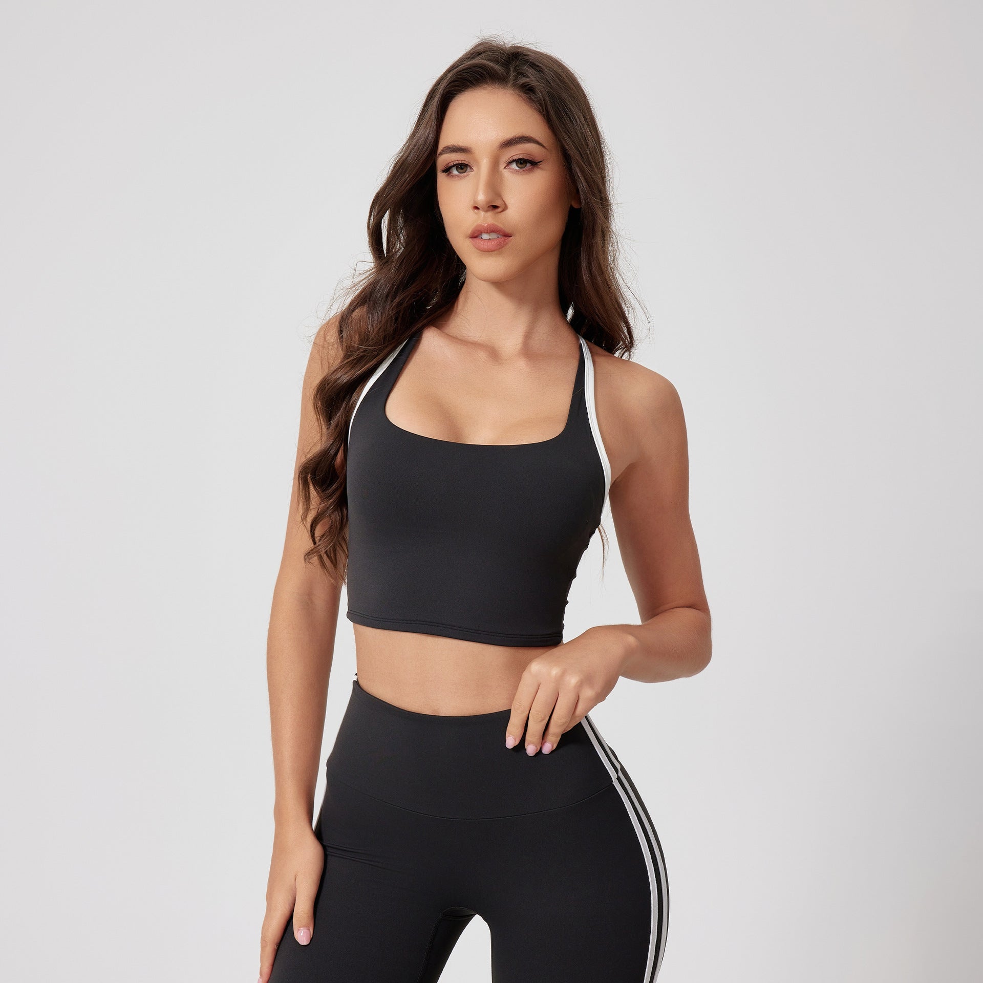 A woman is standing with hand resting on waist in a black racerback sports bra with white trim and matching striped leggings