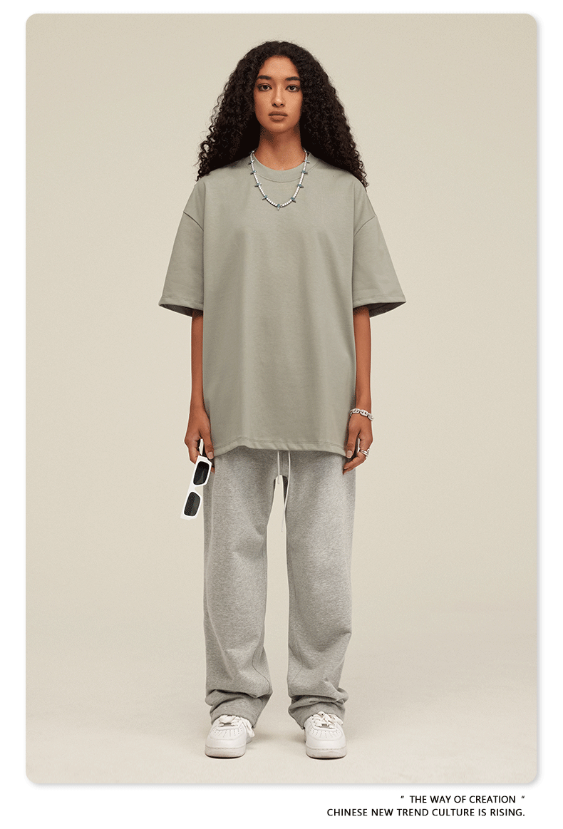 Woman wearing light sage green oversized t-shirt with light gray sweatpants, white sneakers and beaded necklace, holding white sunglasses