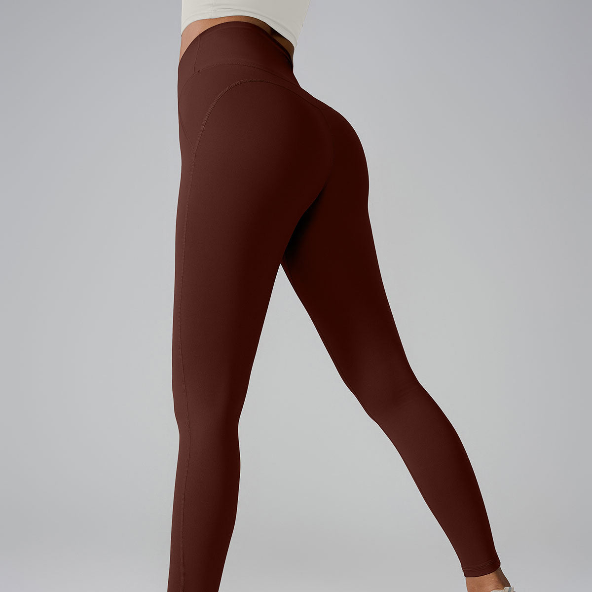 Model in brown high-waisted leggings and cream top, side view showing seamless construction on gray backdrop