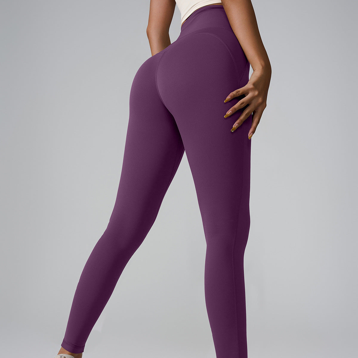 Model in deep purple athletic leggings and cream top, side angle showing seamless design on gray backdrop
