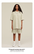 Model with curly hair wearing oversized cream t-shirt and matching shorts with white socks and white sneakers against cream background