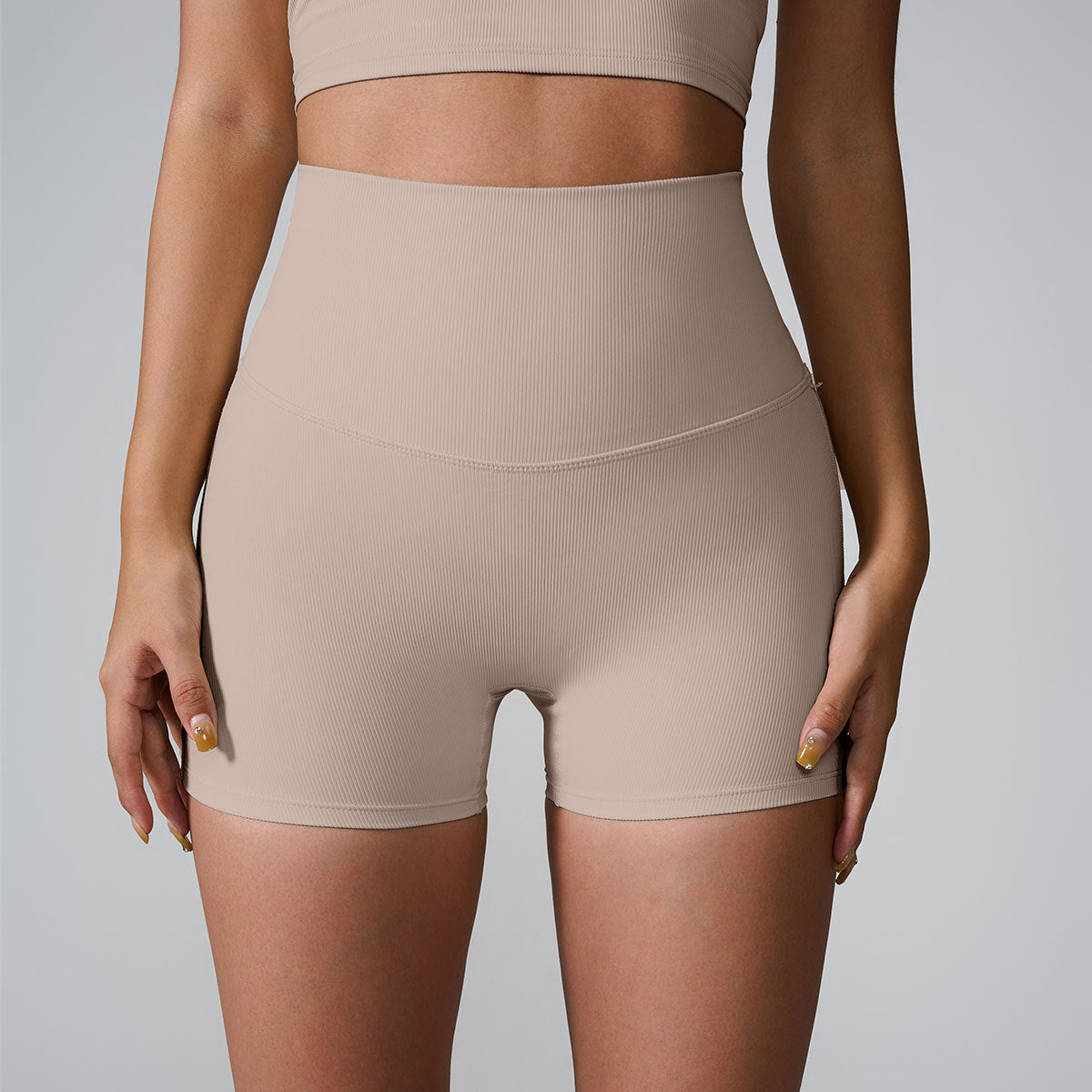 A model wearing beige high-waisted ribbed compression shorts and matching sports bra, photographed against light gray backdrop