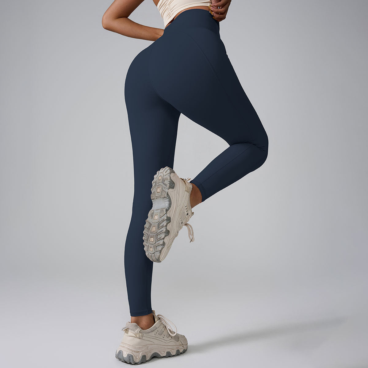 A model wearing navy high-waisted leggings with cream crop top, posing with one leg raised against gray background