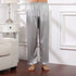 Model wearing light silver silk pajama pants with geometric print and white cuffed hem, standing barefoot on wooden floor next to white nightstand