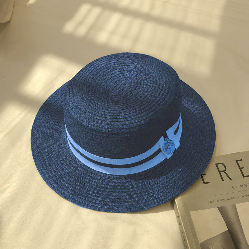 A navy blue straw boater hat with a blue-and-white ribbon band and a silver charm, displayed on a white surface with a magazine nearby.