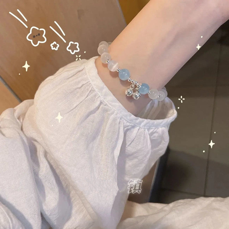 A woman in a white cotton top modeling a gemstone bracelet featuring clear and light blue crystal beads. Silver bell charms dangle from the bracelet