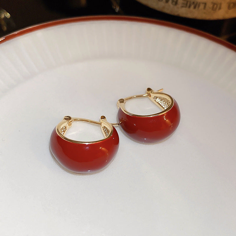 A pair of small red enamel gold hoop earrings with a glossy finish is displayed on a white ceramic plate