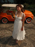 Woman in a white dress standing next to an orange vintage car on a pebbled surface.
