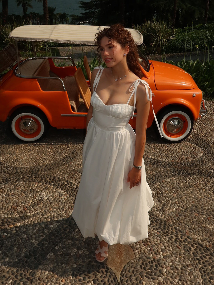 Woman in a white dress standing next to an orange vintage car on a pebbled surface.