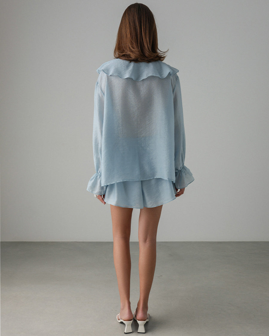 Woman wearing a light blue outfit with ruffled details against a plain background