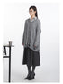 Side view of oversized grey cable-knit sweater featuring V-neckline and distressed bottom edge, paired with charcoal pleated midi skirt and striped collar detail