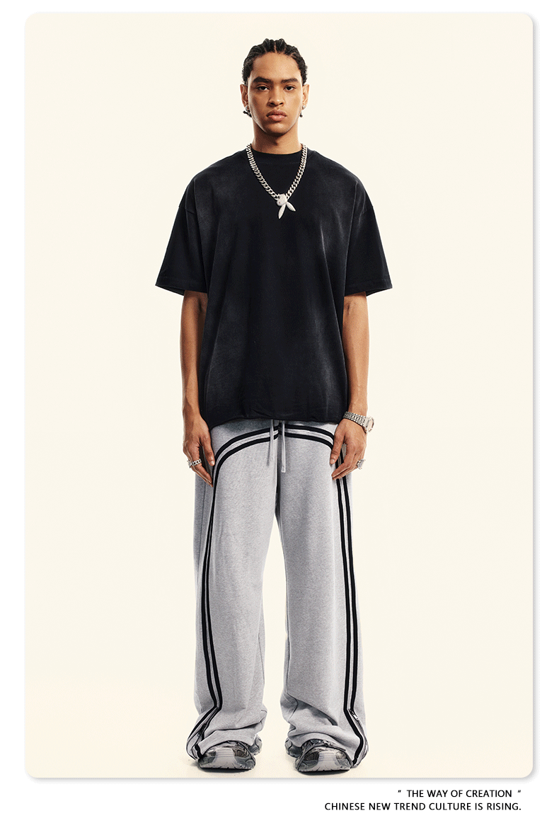 A man wearing oversized black t-shirt and light gray sweatpants with double side stripes stands against neutral background, accessorized with silver chain necklace