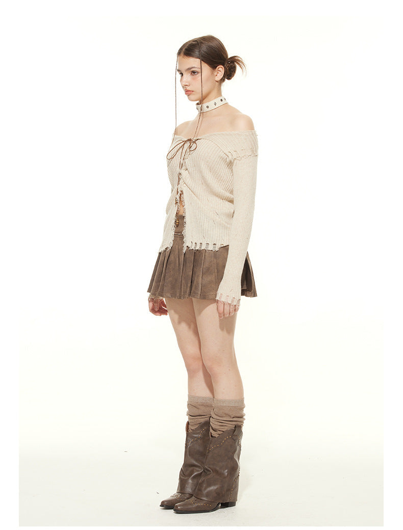Side view of cream ribbed sweater with off-shoulder design, lace-up front detail, and distressed hem. Styled with brown suede pleated skirt, studded choker, and tall brown boots