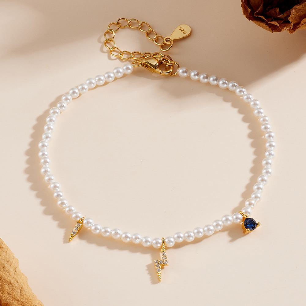 Gold-plated pearl bracelet with lightning bolt and blue heart charms on a beige background.