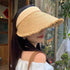 A woman wearing a beige straw visor hat with a soft band and wide frayed brim, paired with a white off-shoulder top for a chic look.