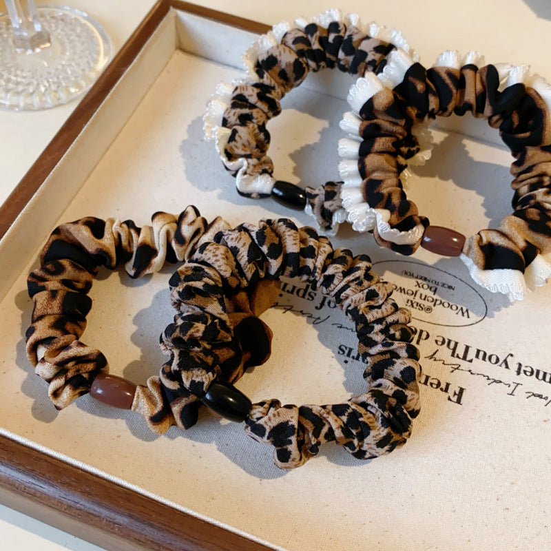 Product flat lay of leopard print scrunchies with white trim and decorative beads displayed on wooden tray against neutral background