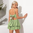 Back view of a woman wearing a green ruffled romper with a bow-tied open back, accessorized with gold earrings, standing in bright sunlight