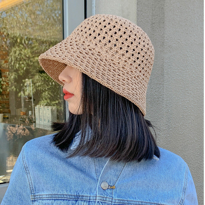 A woman in a khaki open-weave straw bucket hat, wearing a denim jacket and red lipstick, standing in front of a glass building with sunlight filtering through.