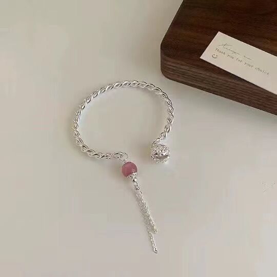 A silver twisted chain bracelet with pink gemstone charm and tassel displayed on white surface with thank you card