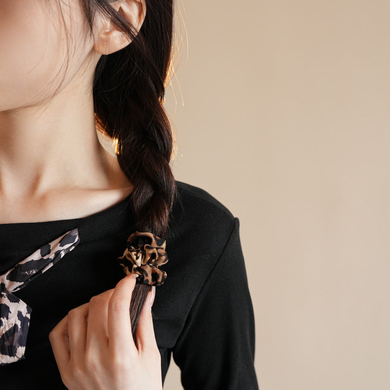 Woman with side braid secured with leopard print scrunchie, hand adjusting hair while wearing black top against beige background