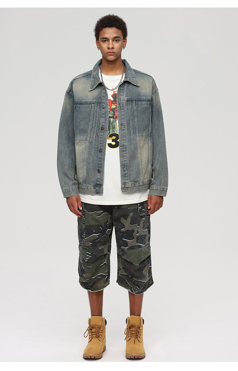 A man is standing front-facing against a gray backdrop. Wearing gray denim jacket, graphic tee with number 3, camouflage cargo shorts and tan work boots