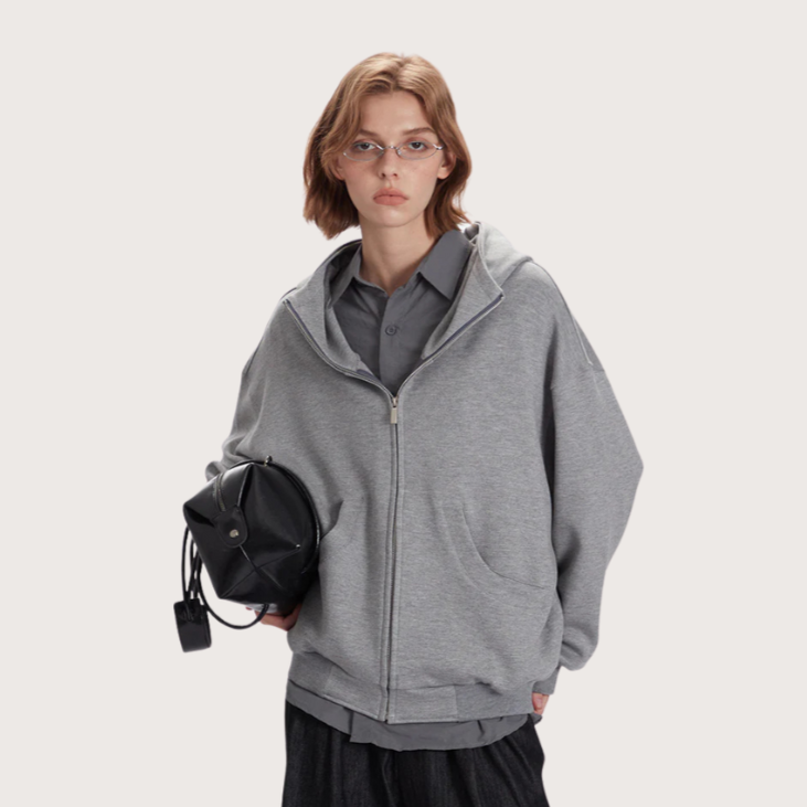Model in oversized gray zip-up hoodie layered over a button-up shirt, styled with dark pants and a black leather bag.