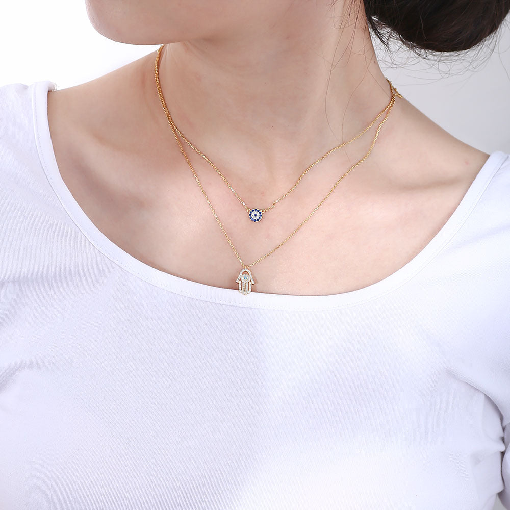 Model wears gold layered necklace with evil eye and Hamsa charms on white top