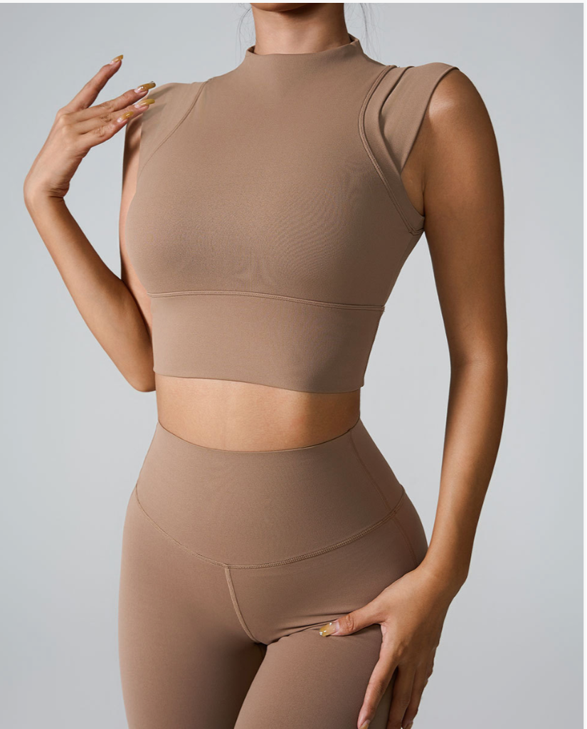 A woman is wearing a nude seamless mock neck crop top with high-waisted leggings, posing with one hand raised against a gray background
