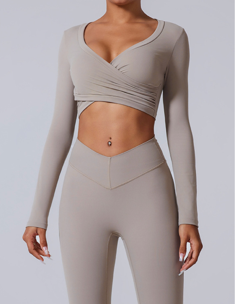 A model wearing a light taupe wrap-style crop top with long sleeves and matching leggings, featuring a V-shaped waistband detail against gray backdrop