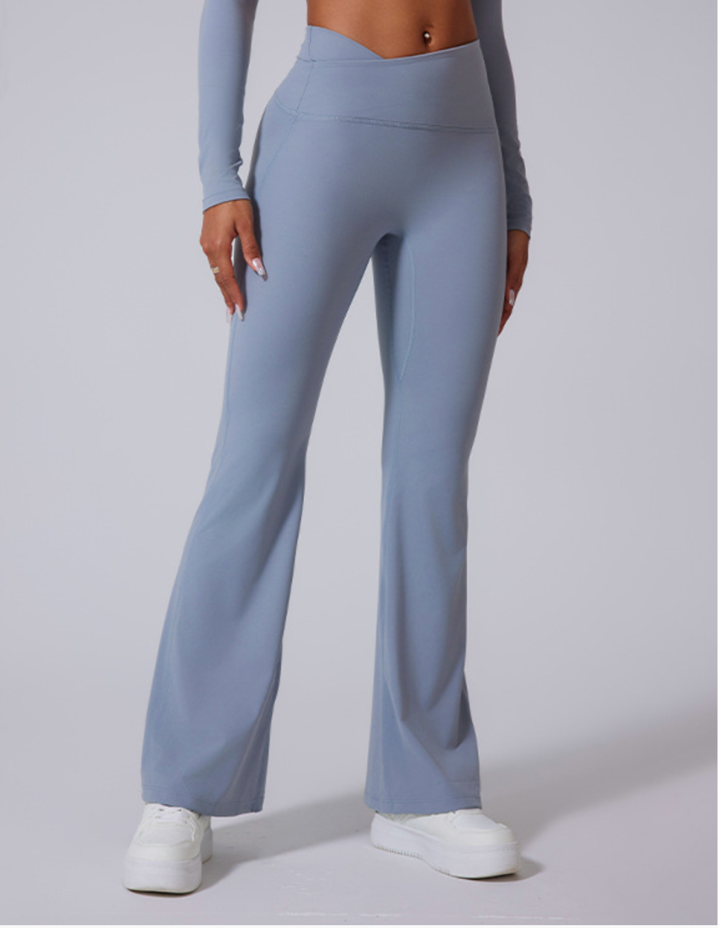 A model wearing powder blue high-waisted flared yoga pants with a seamless design and white platform sneakers on gray background