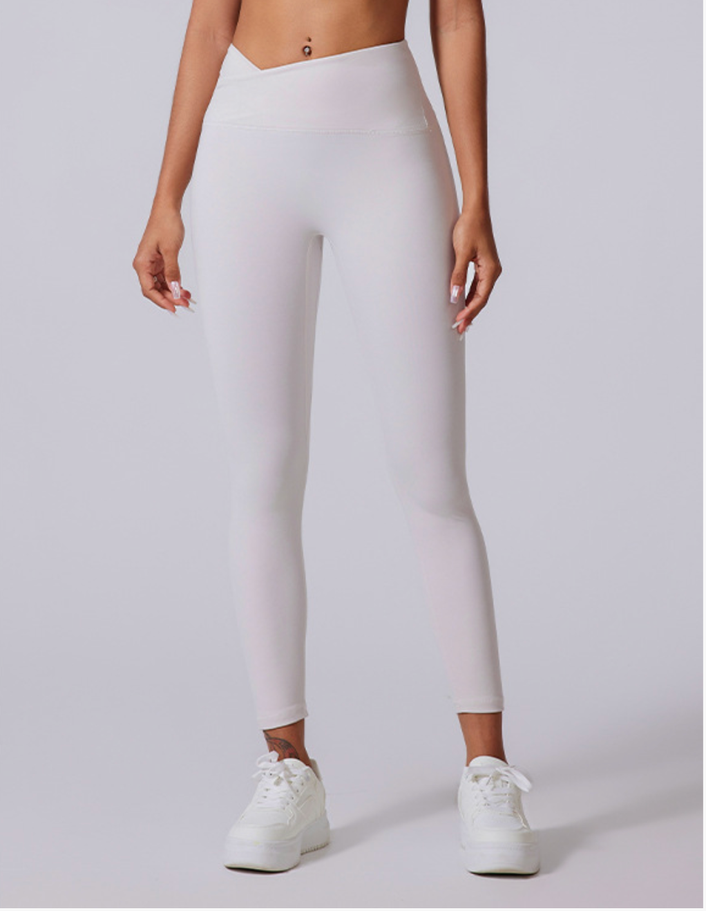 A model is standing wearing white high-waisted leggings with V-shaped waistband paired with white platform sneakers against neutral gray background