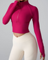 A woman wearing a fuchsia ribbed half-zip crop top with long sleeves and thumb holes paired with cream high-waisted leggings against a gray background