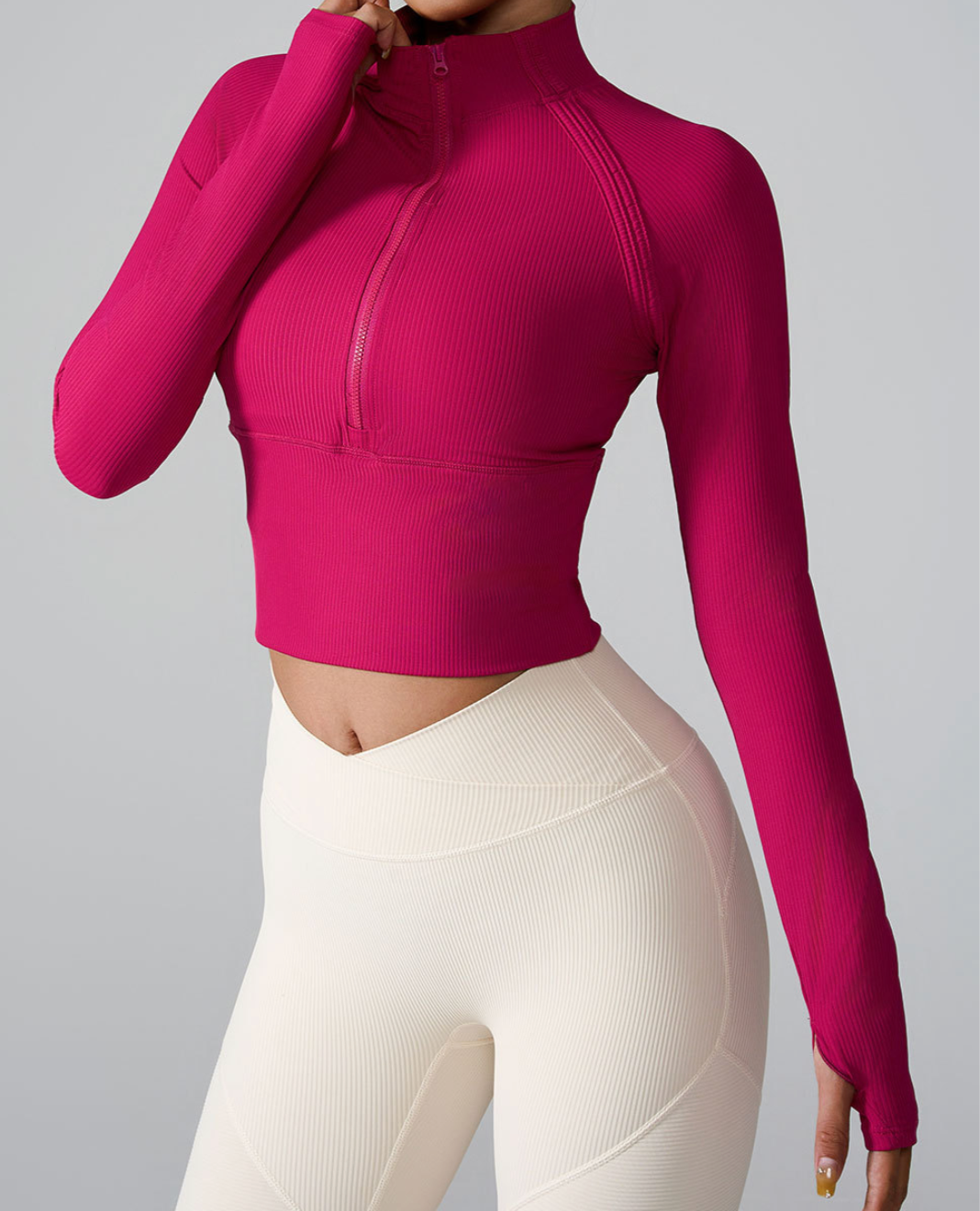 A woman wearing a fuchsia ribbed half-zip crop top with long sleeves and thumb holes paired with cream high-waisted leggings against a gray background