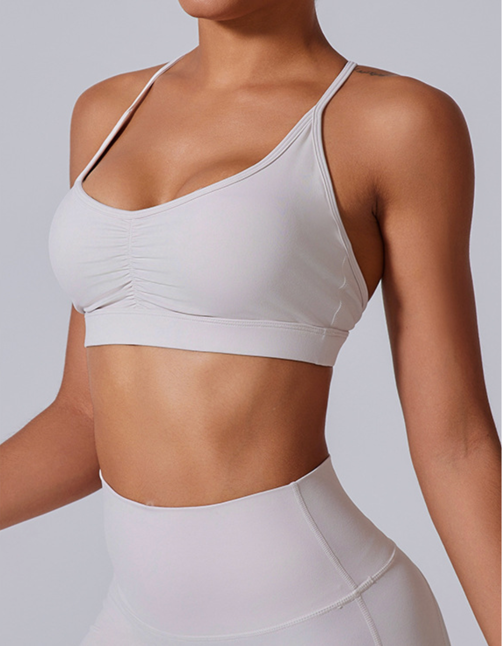 A woman wearing a white strappy sports bra with ruched center detail and matching high-waist leggings against neutral backdrop