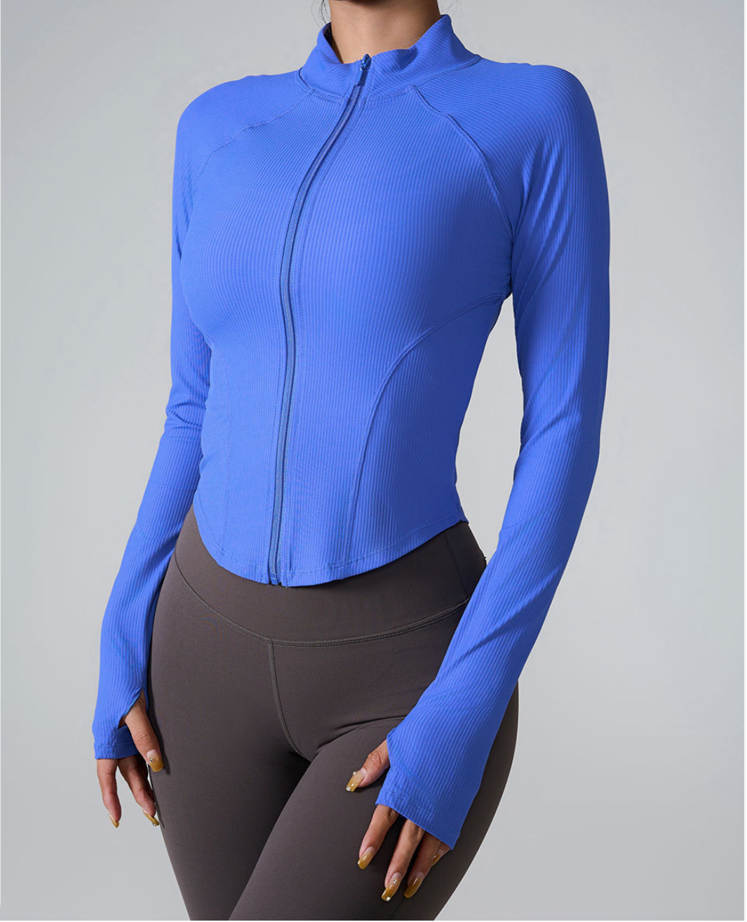 A woman wearing a bright blue ribbed zip-up athletic jacket with raglan sleeves and thumb holes paired with brown high-waisted leggings against a gray background