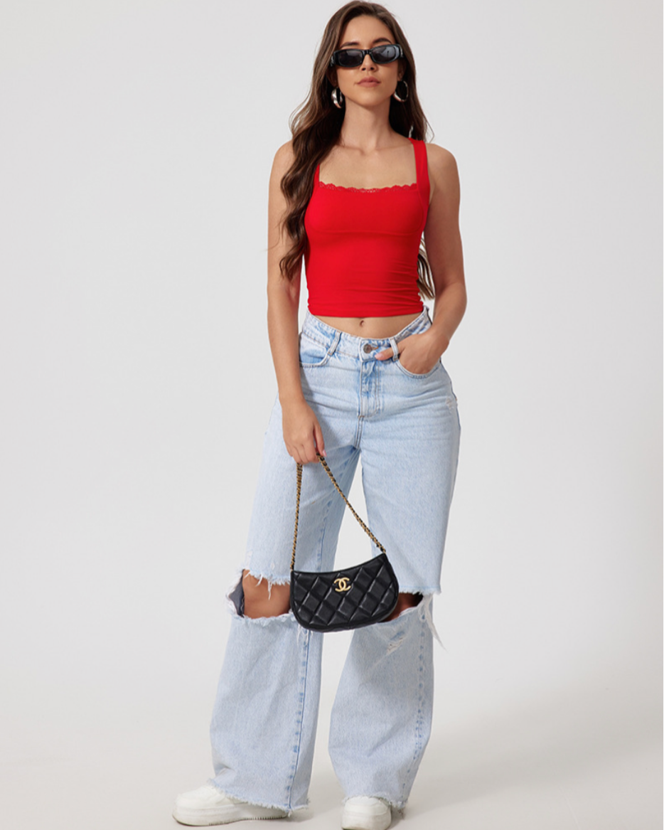 Full body shot of model in red crop top with black sunglasses and ripped light jeans, carrying quilted handbag against white wall