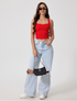 Full body shot of model in red crop top with black sunglasses and ripped light jeans, carrying quilted handbag against white wall