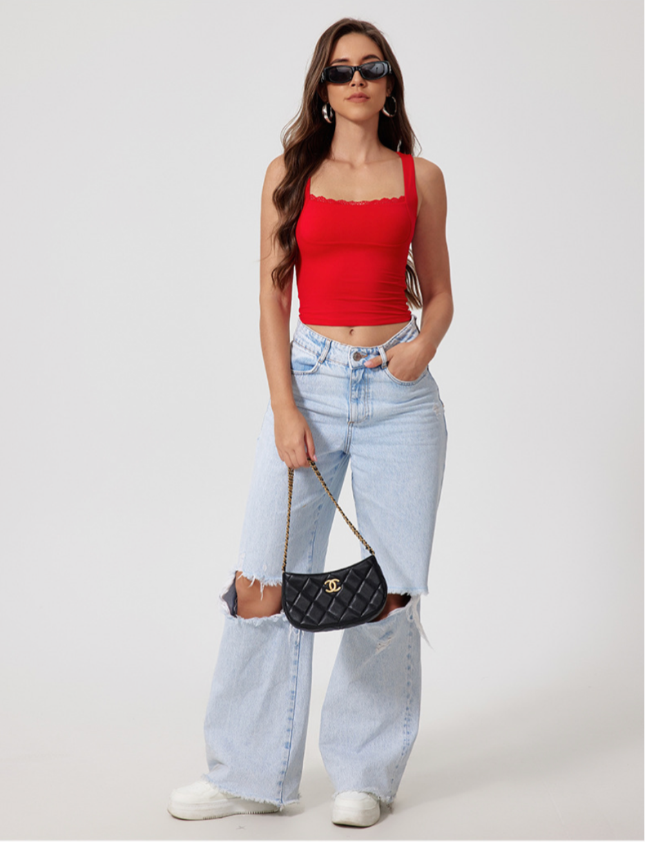 Full body shot of model in red crop top with black sunglasses and ripped light jeans, carrying quilted handbag against white wall