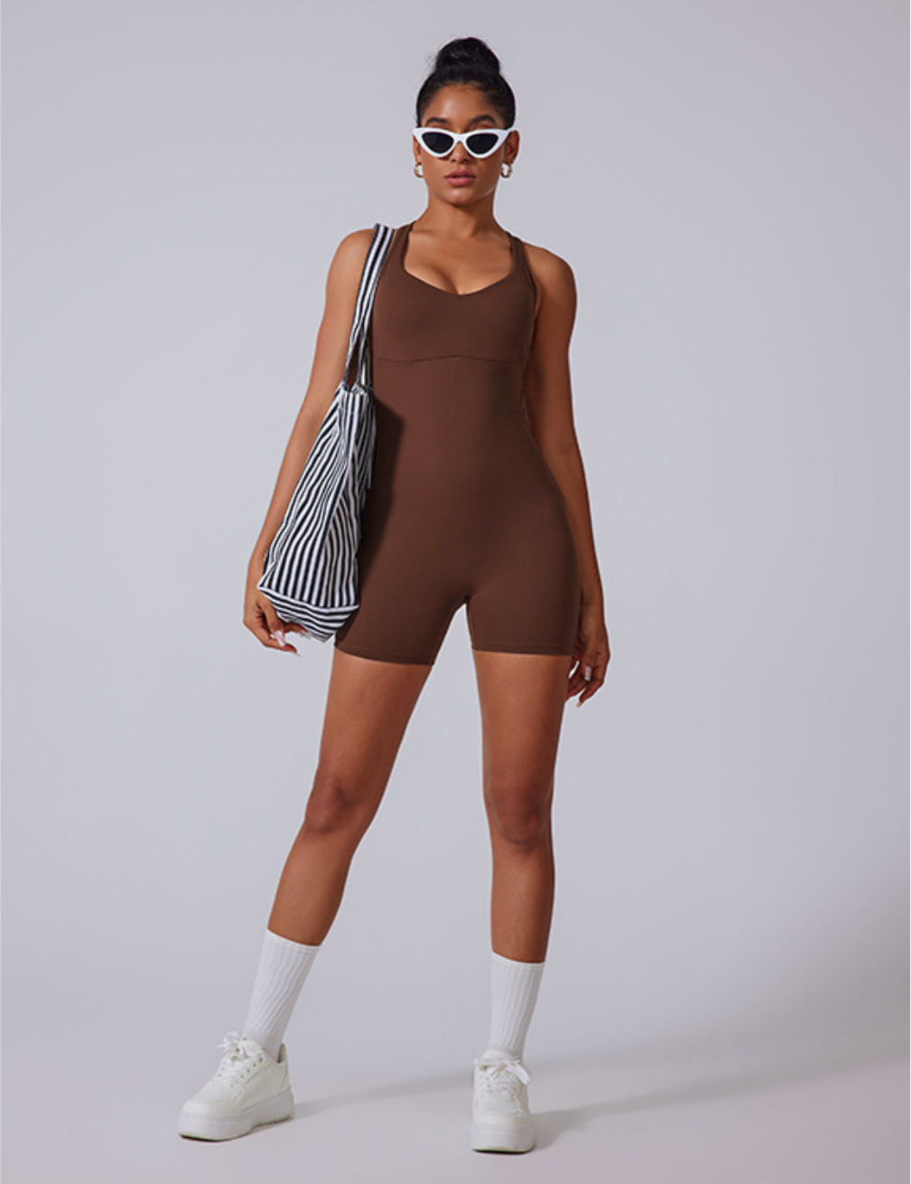 Model wearing chocolate brown sleeveless bodysuit with white cat-eye sunglasses, striped tote bag and white platform sneakers