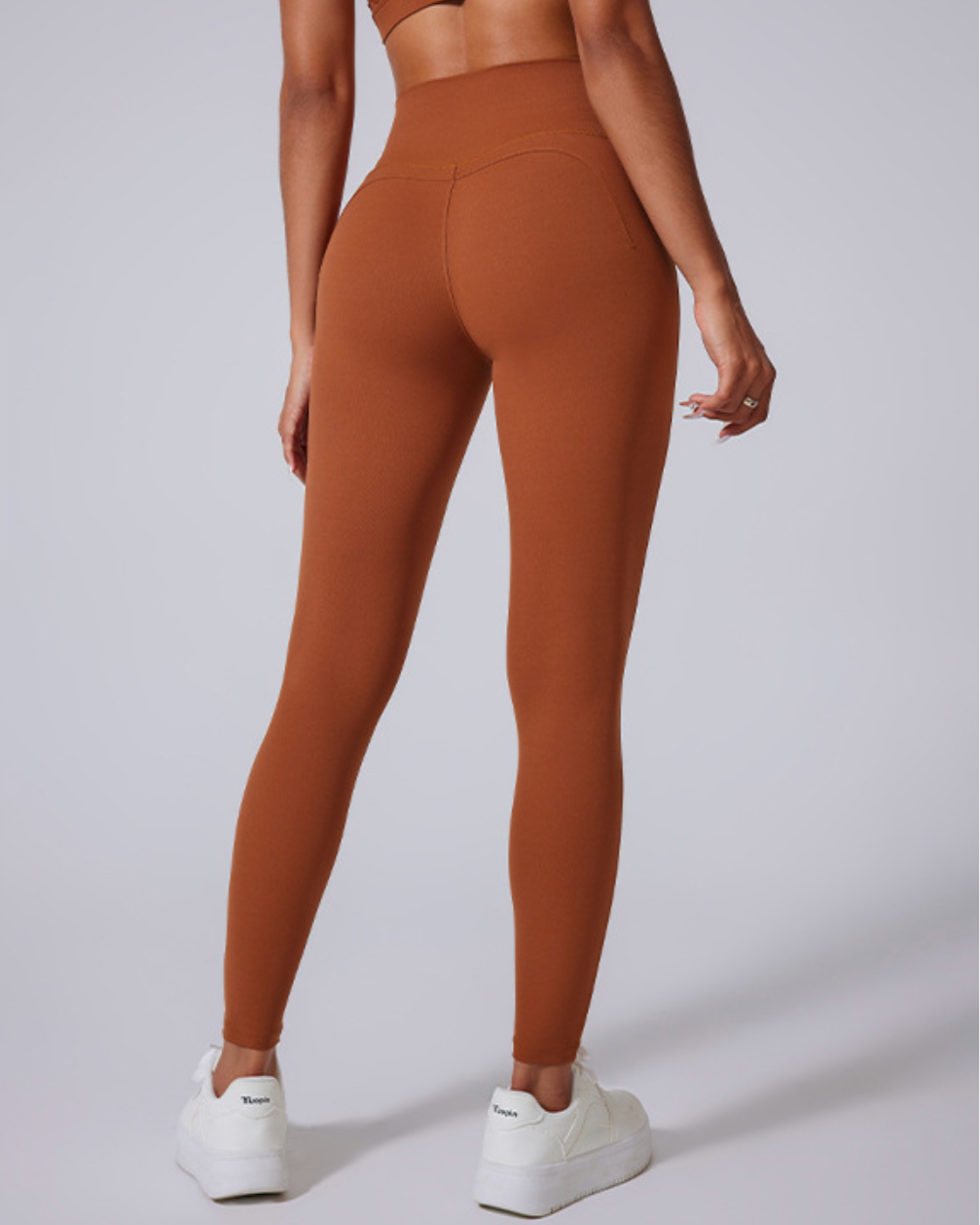 A woman wearing brown high-waisted athletic leggings with curved back seams, photographed from behind against a light gray background with white sneakers