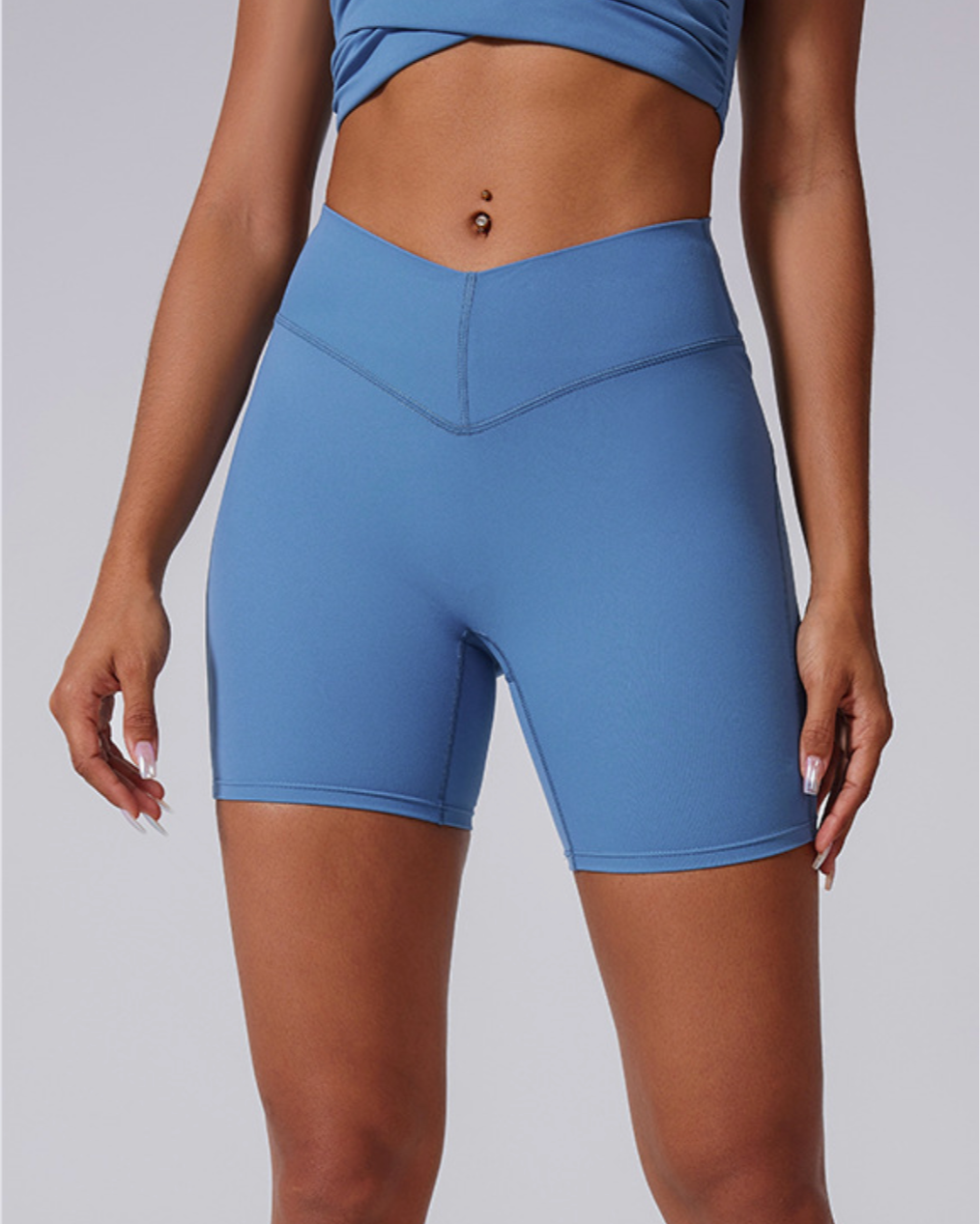 A model is wearing blue high-waisted compression bike shorts with V-shaped waistband and matching sports bra against gray background