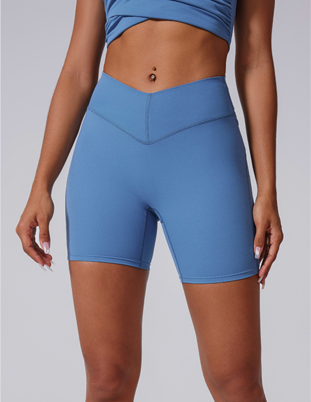 A model is wearing blue high-waisted compression bike shorts with V-shaped waistband and matching sports bra against gray background