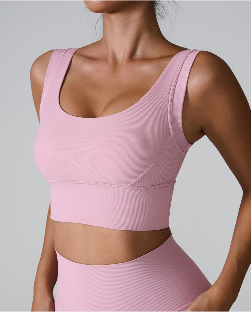A woman wearing a light pink ribbed scoop neck sports bra with wide straps and supportive underbust band paired with matching bottoms against a gray background