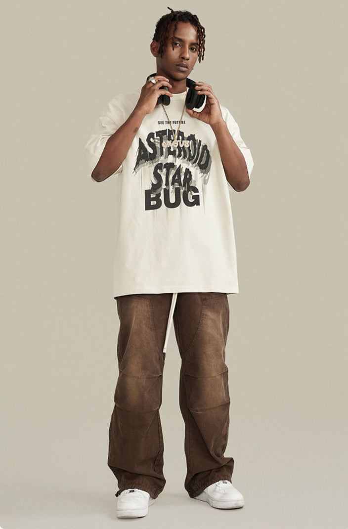 Man in cream graphic tee and brown pants holding headphones around neck.